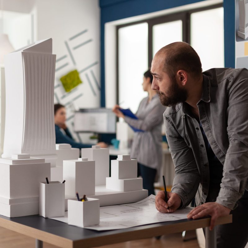 Man working on architectural design at office while looking at laptop computer for industry construction. Project manager inspecting maquette modern design building model